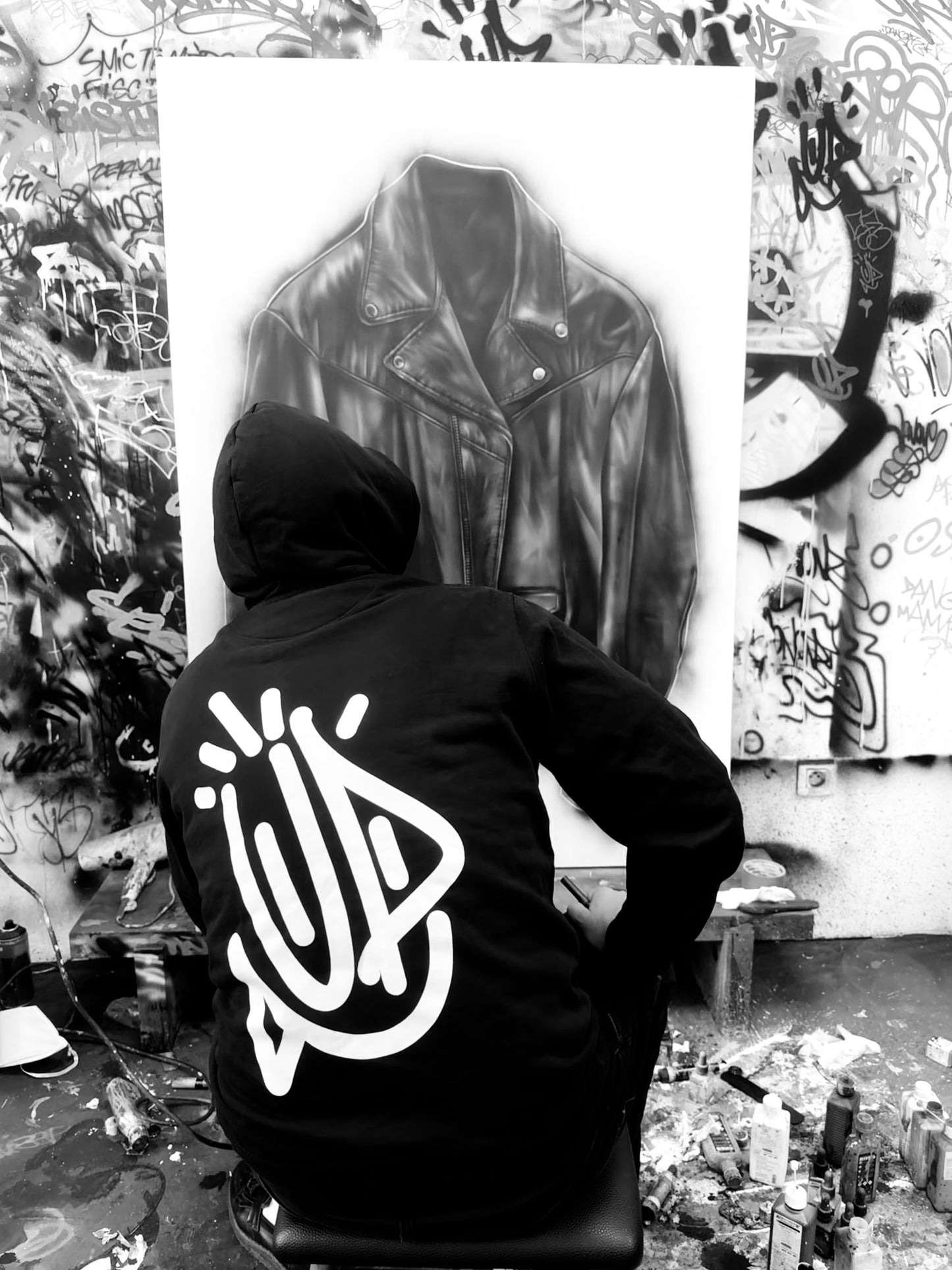 Artiste Onemizer de dos dans son atelier, portant le sweat 4U avec le large graffiti signature blanc sur fond de murs tagués et bombes de peinture.